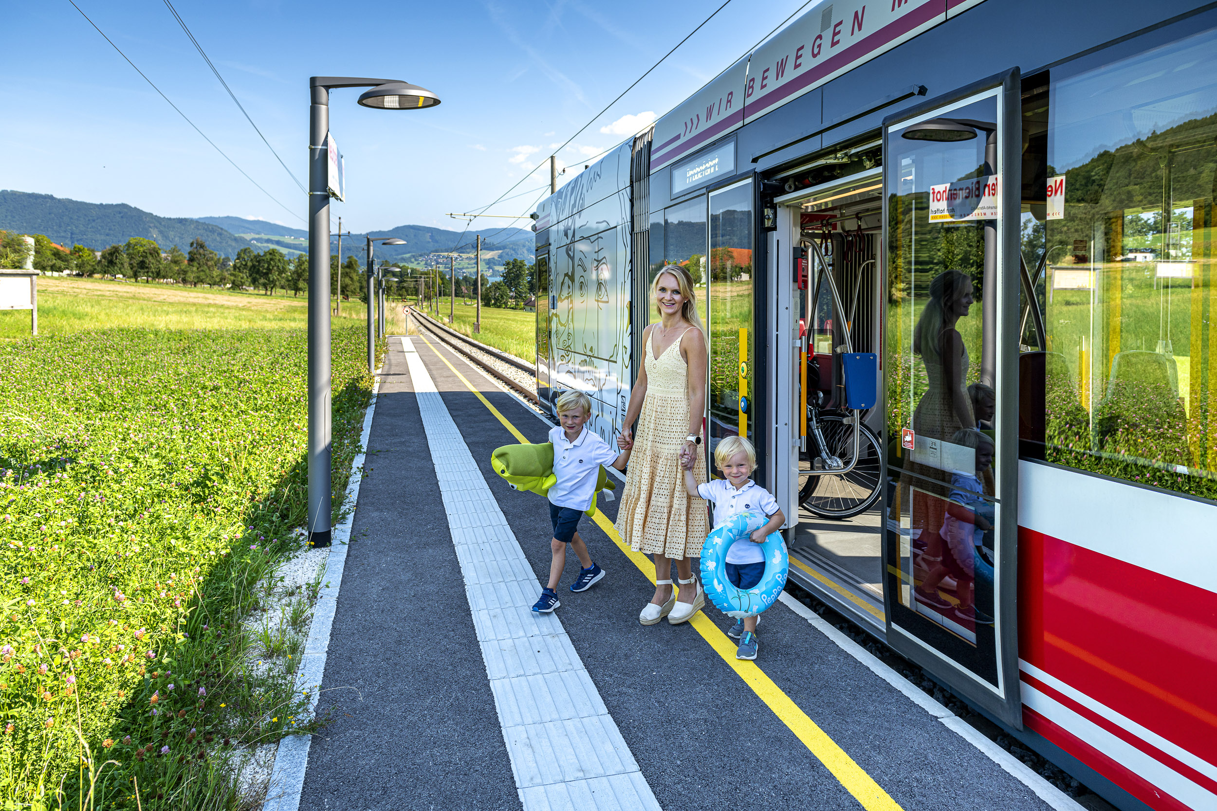 eine Frau mit ihren 2 Söhnen steigt mit Badespielzeug aus der Atterseebahn aus